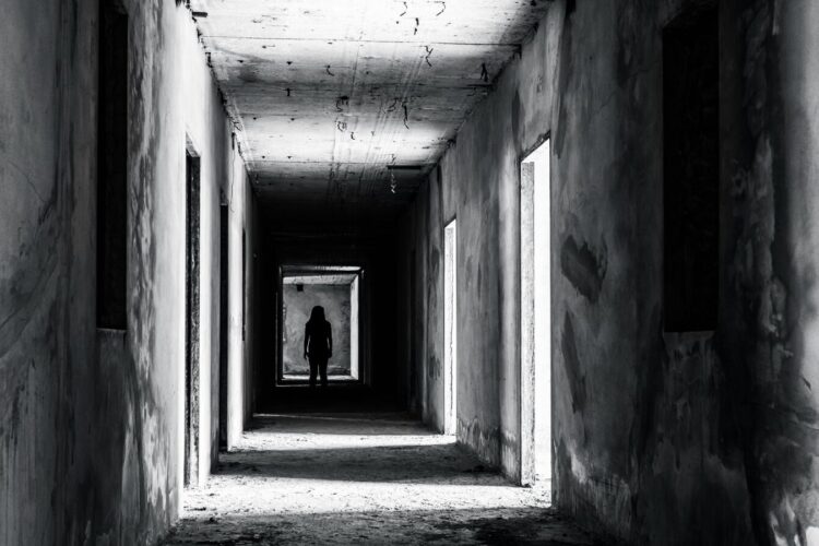 A person stands in silhouette at the end of a dark, empty corridor in a dilapidated building with visible damage and open doorways.