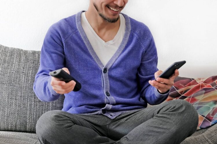 Man sitting cross-legged on a couch, holding a TV remote in one hand and a smartphone in the other, smiling.