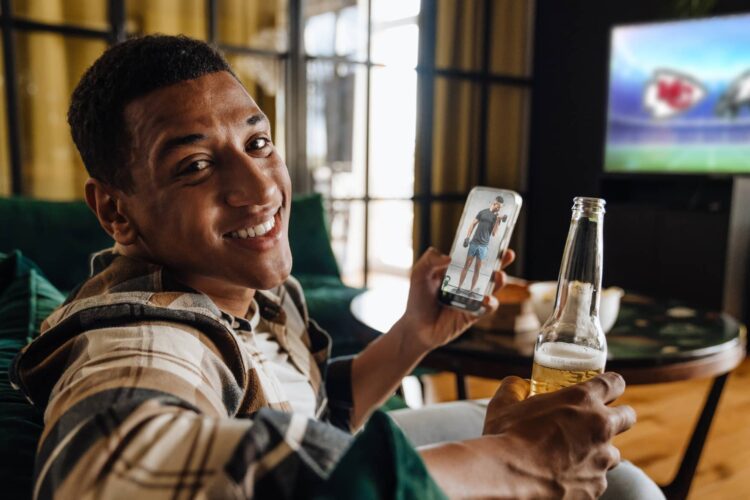 Man sitting on a couch holding a beer and a smartphone, smiling at the camera, with a football game on a TV in the background.