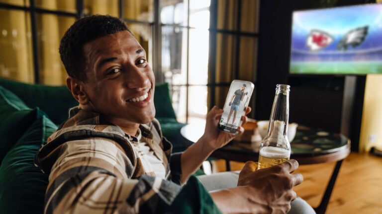 Man sitting on a couch holding a beer and a smartphone, smiling at the camera, with a football game on a TV in the background.