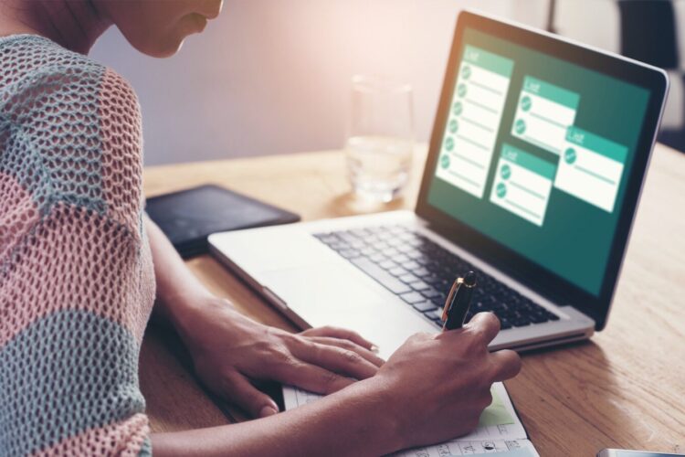 Person writes in a notebook while looking at a laptop displaying a task management or calendar application on a wooden desk.