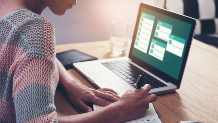Person writes in a notebook while looking at a laptop displaying a task management or calendar application on a wooden desk.
