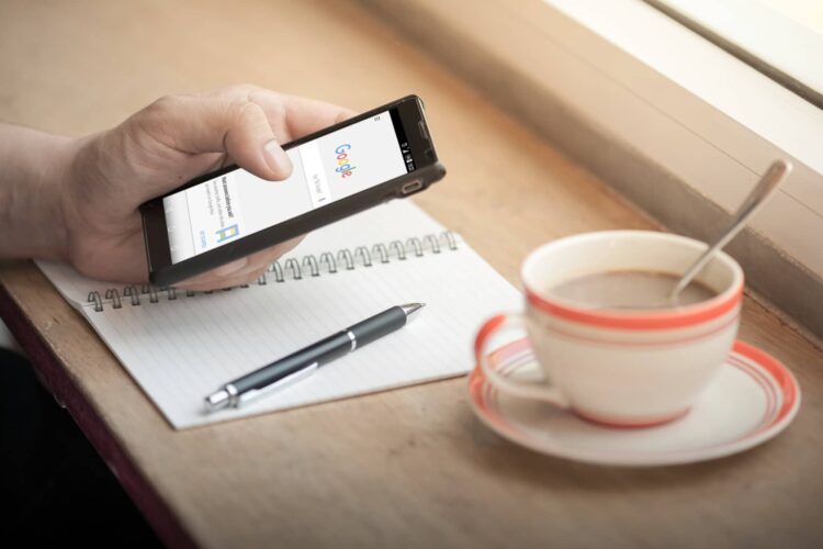 A person holds a smartphone displaying Google search next to a notepad, pen, and a cup of coffee on a wooden table by a window.