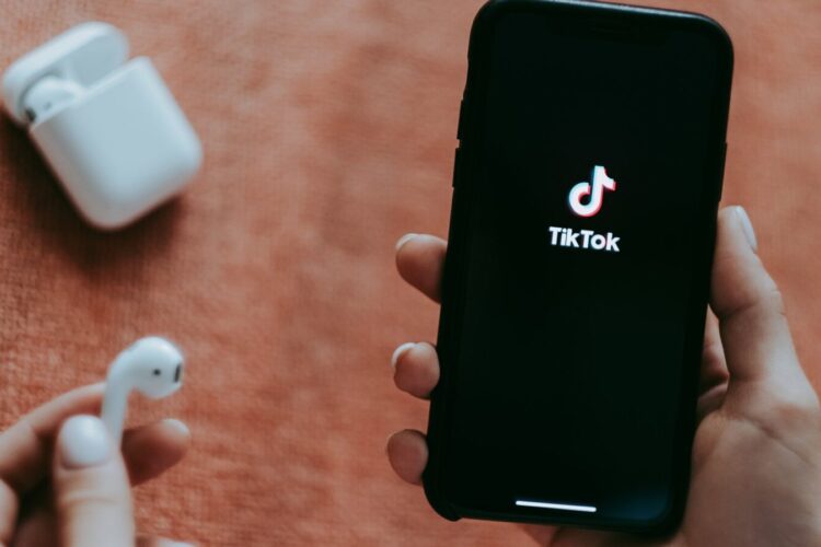 A person holds a smartphone displaying the TikTok app logo, with an open wireless earbud case and one earbud visible on an orange surface.