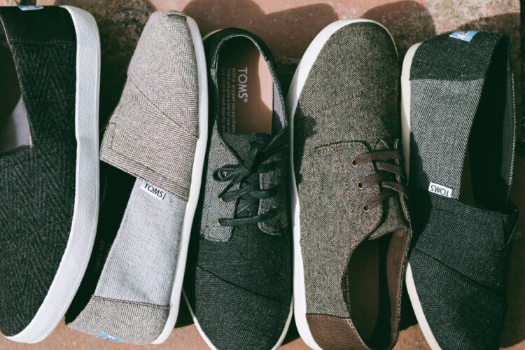Five pairs of TOMS shoes in various neutral colors and styles are lined up side by side on a stone surface.