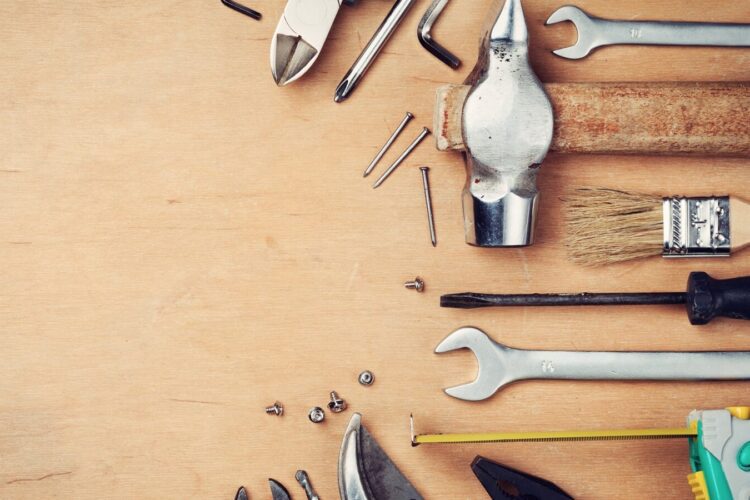 Various hand tools, including a hammer, wrenches, pliers, screwdriver, paintbrush, and measuring tape, arranged on a wooden surface.