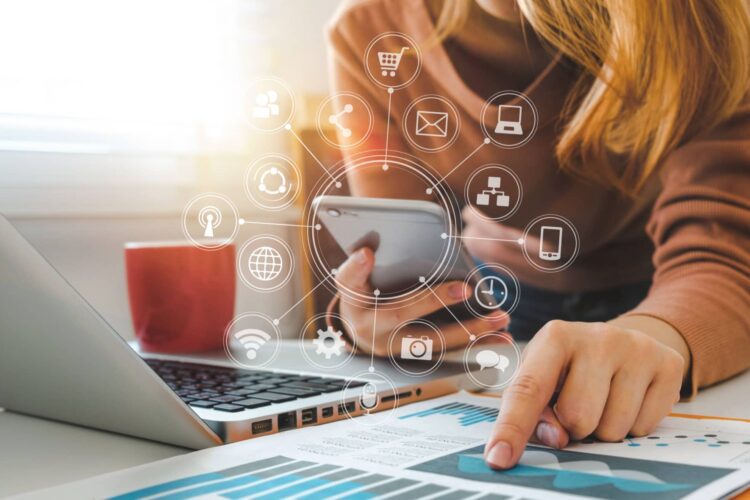 Person using a smartphone and pointing at charts on paper beside a laptop, with digital icons representing various online services and apps overlaid on the image.