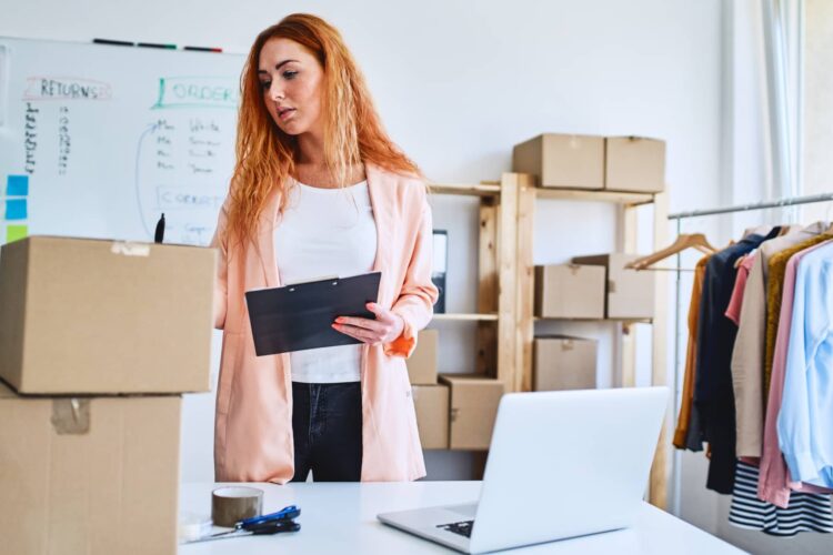 A woman stands in an office with boxes, holding a clipboard and pen, next to a laptop. Shelves with packages and clothes are visible in the background.