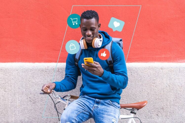 A man in a blue hoodie sits on a bicycle, looking at his phone, with social media icons digitally displayed around him against a red wall.