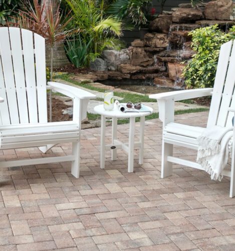 Two white Adirondack chairs with a small round table between them on a brick patio, beside a garden with a waterfall and plants.
