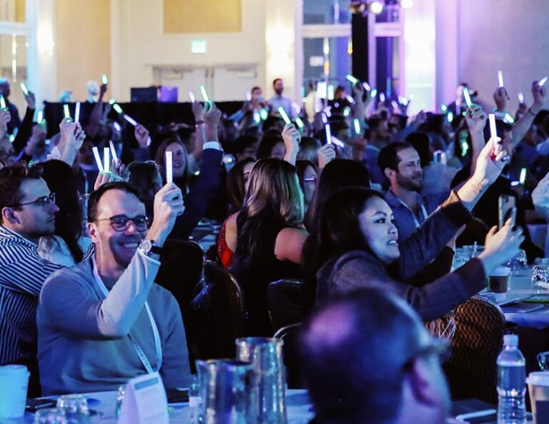A seated audience holds up glow sticks in a conference room, with many people smiling and looking toward the stage.