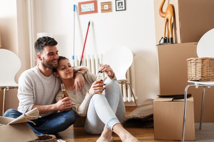 A couple sits on the floor among moving boxes, smiling and holding drinks, with unpacked items and chairs around them in a bright room.