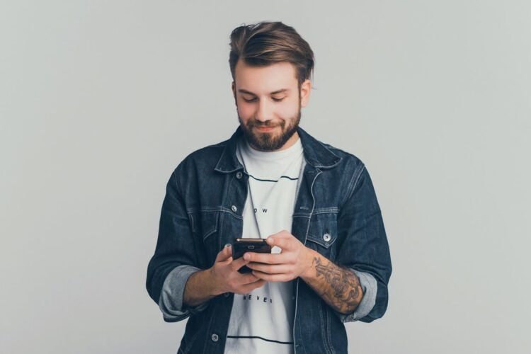 A man with a beard and tattooed arm is standing against a plain background, looking down and smiling while using a smartphone.
