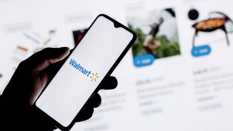 A hand holds a smartphone displaying the Walmart logo, with a blurred online shopping website shown in the background.