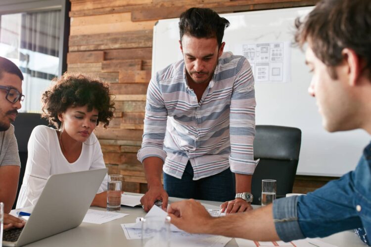 Four people sit and stand around a table in an office, discussing documents and a laptop, with papers and glasses of water visible. A whiteboard is in the background.