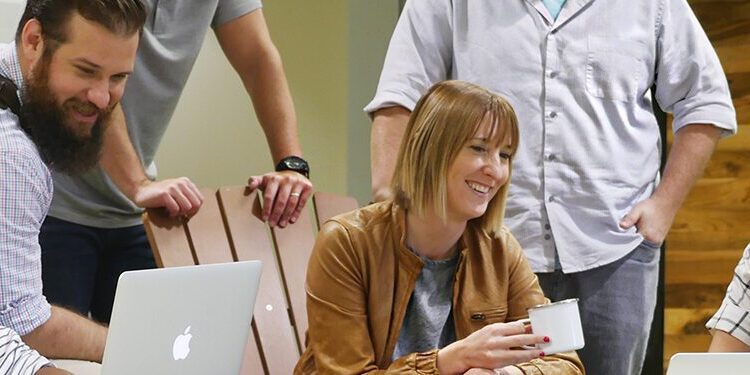 Five people sit and stand around laptops in a casual office, collaborating and smiling during a meeting.