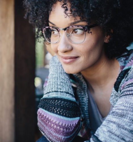 A woman with curly hair and glasses, wearing a striped sweater, looks to the side with a slight smile while leaning on her arms.