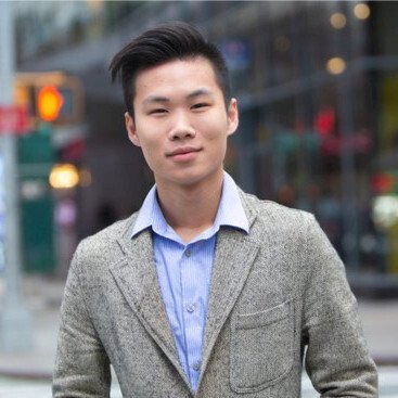 A young man in a gray blazer and blue shirt stands outdoors in an urban setting, with blurred buildings and traffic lights in the background.