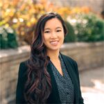 A woman with long, wavy hair wearing a black blazer and dotted shirt stands outdoors in front of a stone wall and blurred foliage, smiling at the camera.