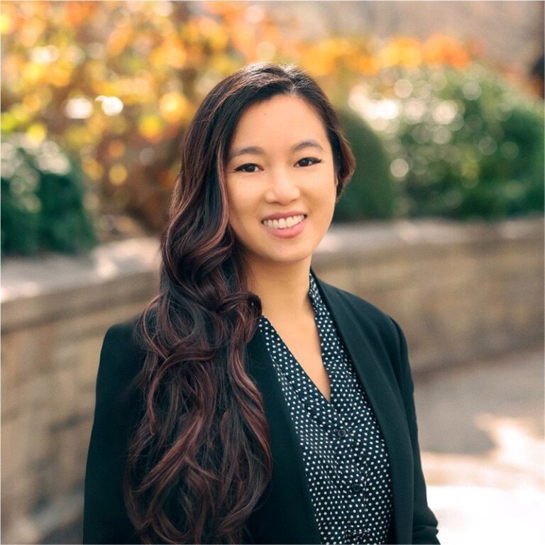 A woman with long, wavy hair wearing a black blazer and dotted shirt stands outdoors in front of a stone wall and blurred foliage, smiling at the camera.