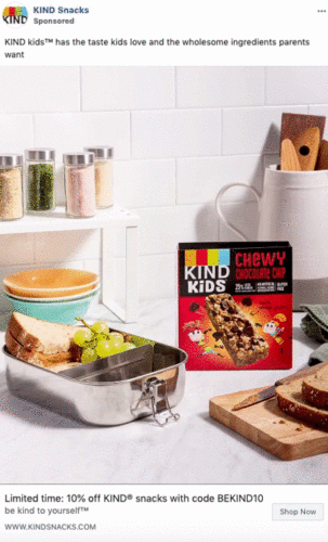A KIND Kids Chewy Chocolate Chip granola bar box is on a kitchen counter next to a lunchbox with a sandwich and grapes, with jars of spices and kitchen utensils in the background.