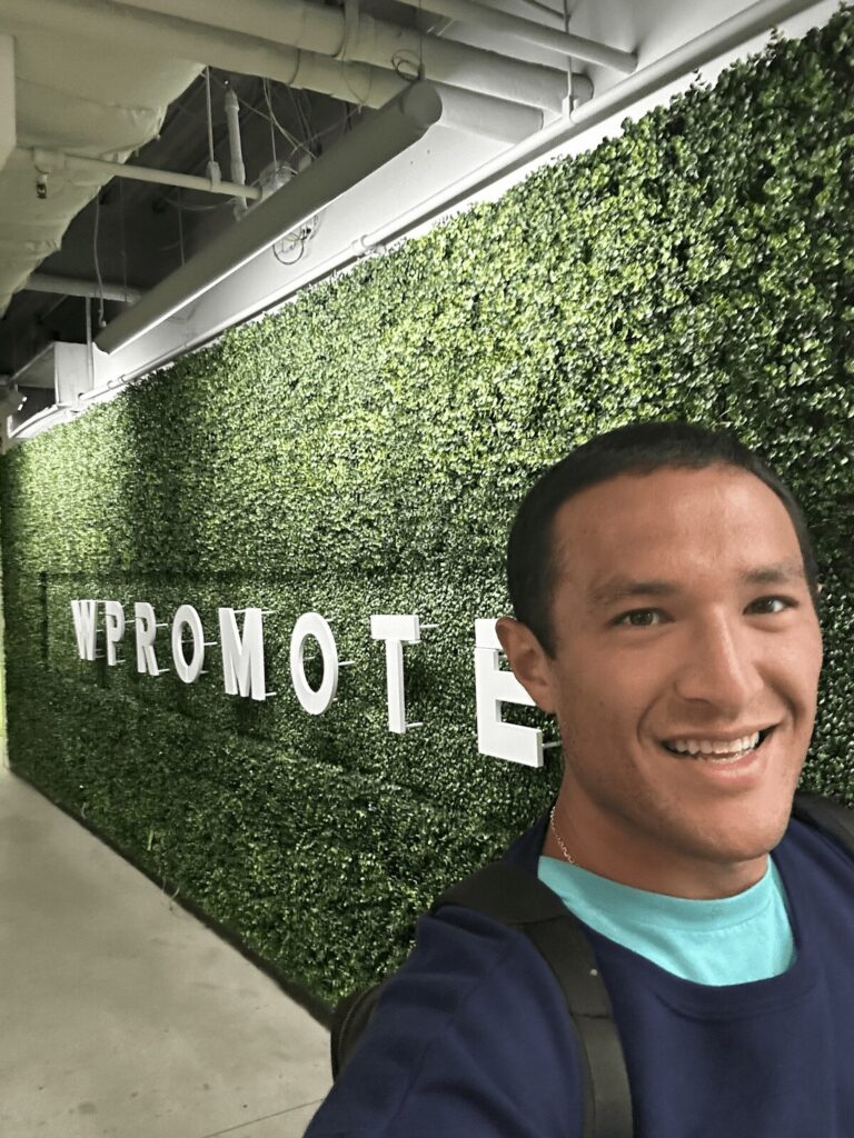 A person smiling in front of a green hedge wall with large white letters spelling "WPROMOTE" in an indoor hallway.