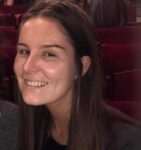 A young woman with long brown hair smiles at the camera while sitting in a theater with red seats.