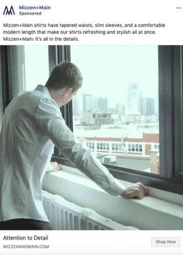 A man in a light blue shirt stands indoors, looking out of a window at a cityscape with tall buildings in the background.