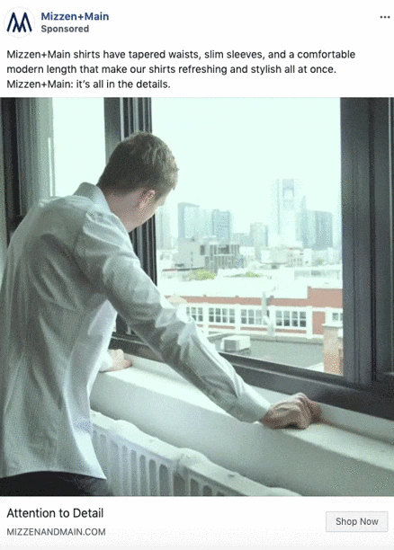 A man in a light blue shirt stands indoors, looking out of a window at a cityscape with tall buildings in the background.