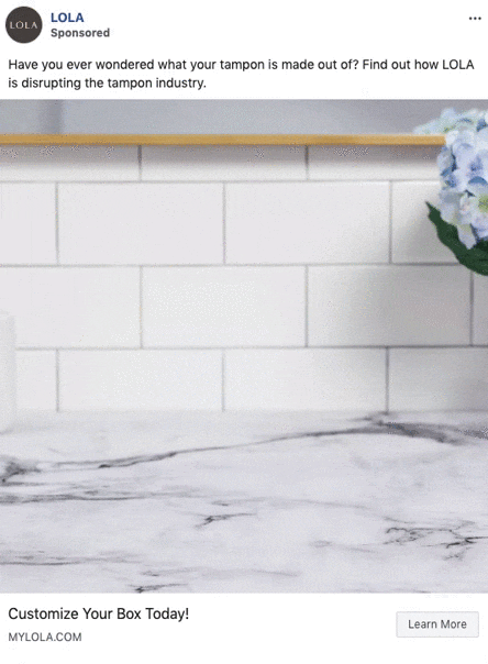 A hand places a LOLA tampon box on a marble countertop in a white-tiled kitchen, with blue flowers in the background.