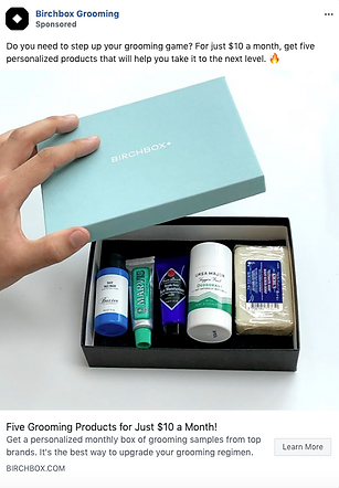 A hand opens a Birchbox containing five assorted grooming products, including tubes, a jar, and a bar of soap, displayed in a black box with a light blue lid.