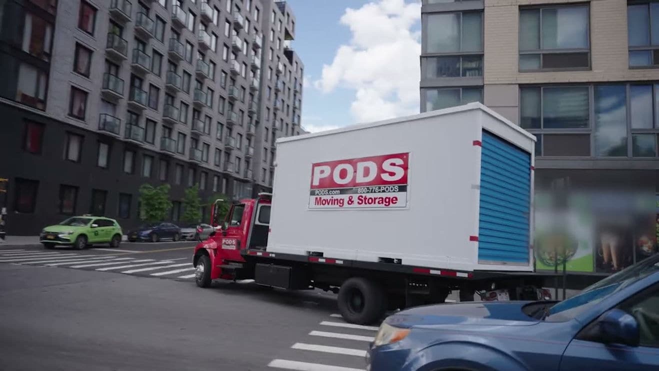 A red truck carrying a white PODS moving and storage container drives through an urban intersection lined with apartment buildings.