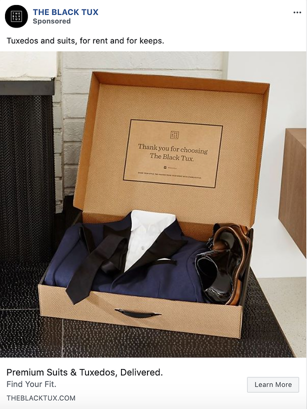 An open box with a suit jacket, white shirt, black tie, and black dress shoes neatly arranged inside, placed on a bench next to a brick wall.