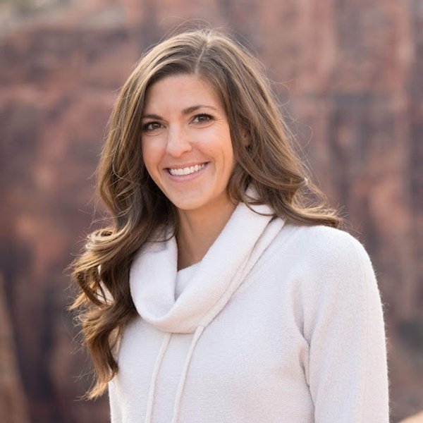 A woman with long brown hair wearing a light-colored sweater stands outdoors in front of a blurred rocky background, smiling at the camera.