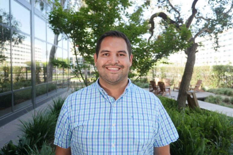 A man wearing a blue and green plaid shirt smiles at the camera while standing outdoors near trees and a building with glass windows.