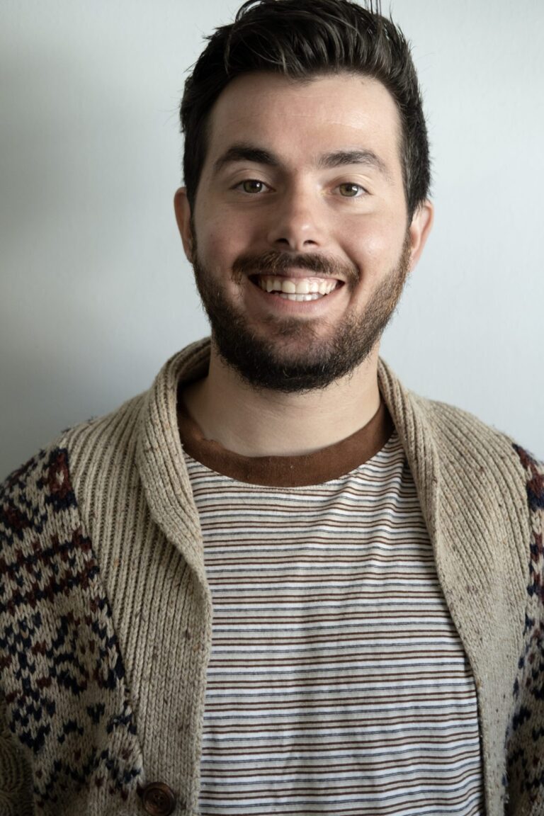 A man with dark hair and a beard smiles at the camera. He is wearing a patterned cardigan over a striped shirt, standing in front of a plain light-colored wall.