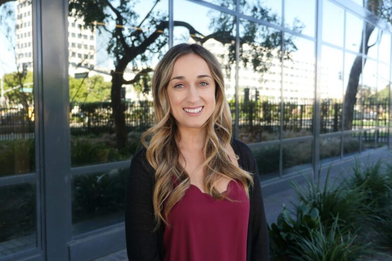 A woman with long, wavy blonde hair wearing a burgundy top and black cardigan stands outside in front of a glass building reflecting trees and nearby structures.