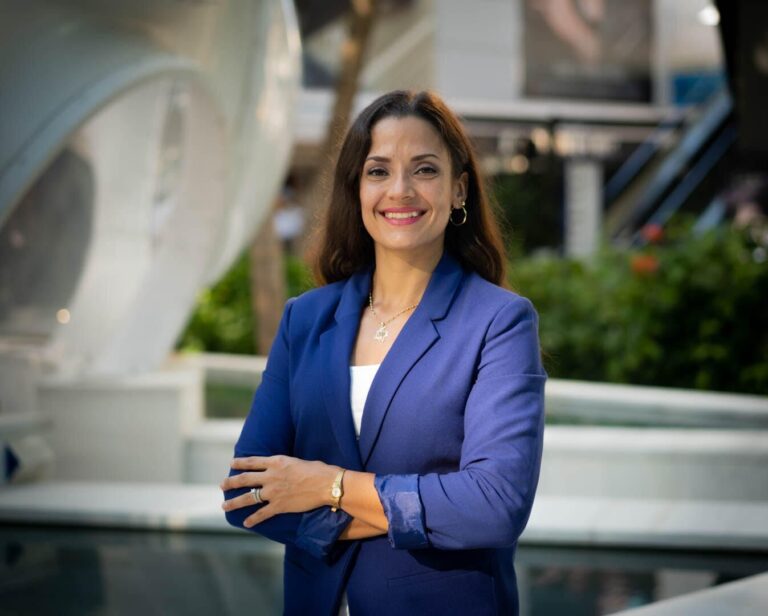 A woman with long brown hair, wearing a blue blazer and white top, stands outdoors with her arms crossed, smiling at the camera. There is greenery and modern architecture in the background.