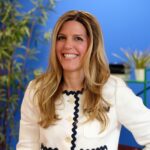 A woman with long blonde hair wearing a white jacket with black trim stands smiling in front of a blue wall with green plants in the background.