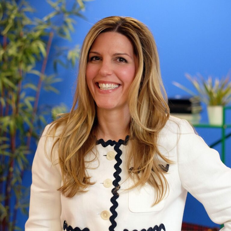 A woman with long blonde hair wearing a white jacket with black trim stands smiling in front of a blue wall with green plants in the background.