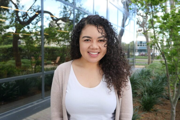 A woman with curly hair, wearing a white shirt and beige cardigan, stands outside in front of a glass building surrounded by greenery.
