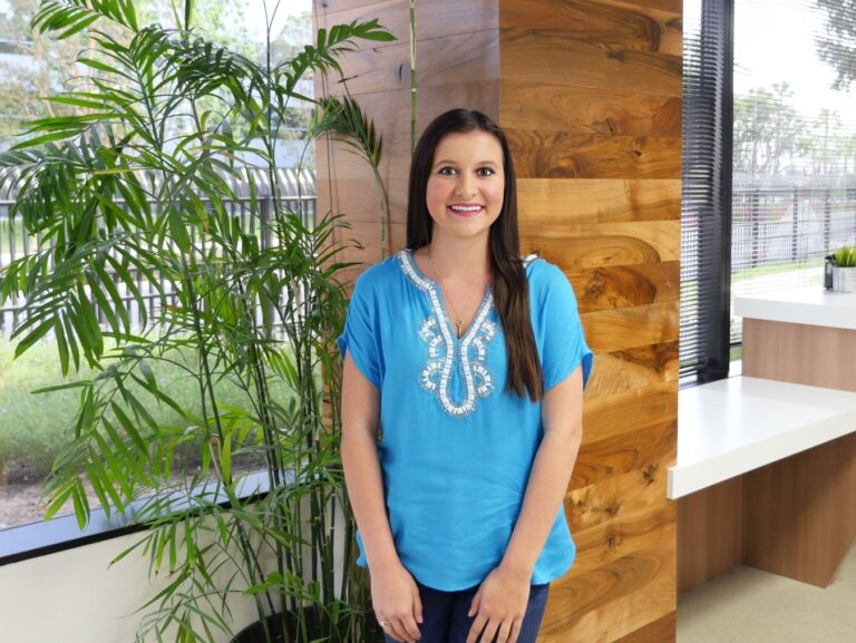 A woman with long brown hair, wearing a blue embroidered blouse, stands indoors next to a tall green plant and wooden wall, with windows in the background.