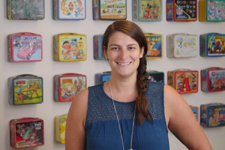 A woman with brown hair in a side braid stands smiling in front of a wall display of colorful vintage lunchboxes.