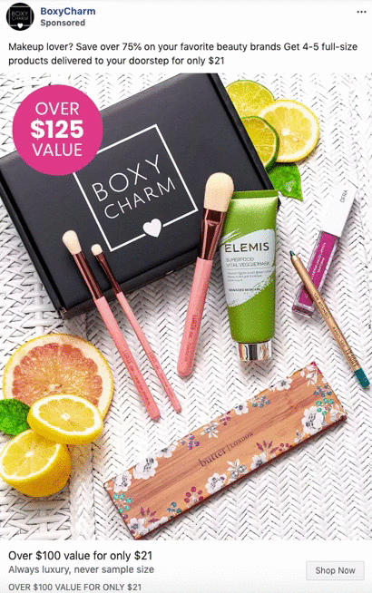 BoxyCharm box with makeup brushes, palette, pencil, ELEMI face wash, and sliced citrus arranged on a white woven surface. Text highlights over $125 value for $21.