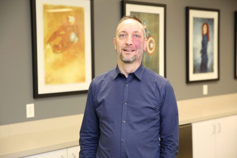 A man in a blue checkered shirt stands indoors in front of framed superhero artwork on a wall.