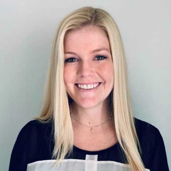 A woman with straight long blonde hair and a black top is smiling at the camera against a light-colored background.
