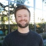 A man with short brown hair and a beard smiles at the camera, standing outside in front of a reflective glass building.