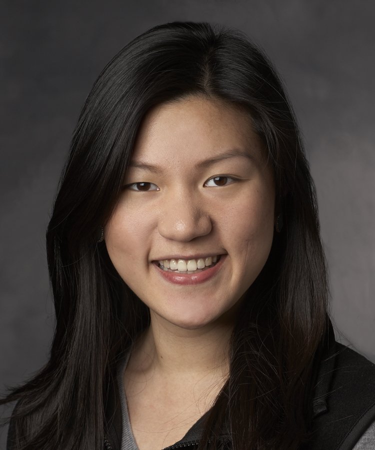 A woman with long, straight black hair smiles at the camera in a professional headshot against a dark, neutral background.