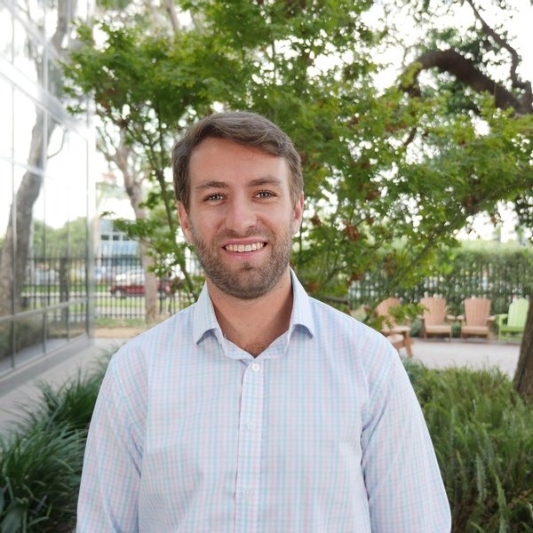 A man with short brown hair and a beard, wearing a light-colored checkered shirt, stands outdoors in front of trees and greenery, smiling at the camera.