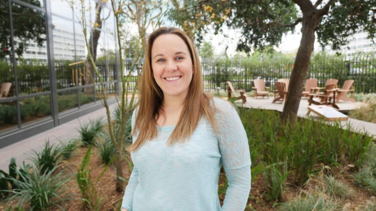 A woman with long brown hair stands outdoors in a landscaped garden area, smiling at the camera; patio chairs and trees are visible in the background.